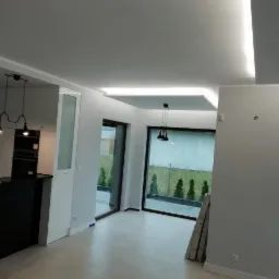 Nowoczesne wnętrze z oświetleniem LED w suficie podwieszanym, widoczne gładkie ściany, duże okna z widokiem na ogród i fragment zabudowy kuchennej z czarnym blatem.