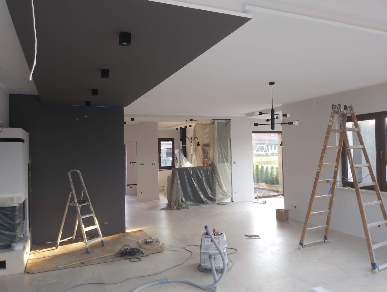 Wnętrze w trakcie remontu: pomalowany na szaro fragment sufitu i ściany, widoczne drabiny, odkurzacz przemysłowy i zabezpieczona folią kuchnia, surowy stan wykończenia.
