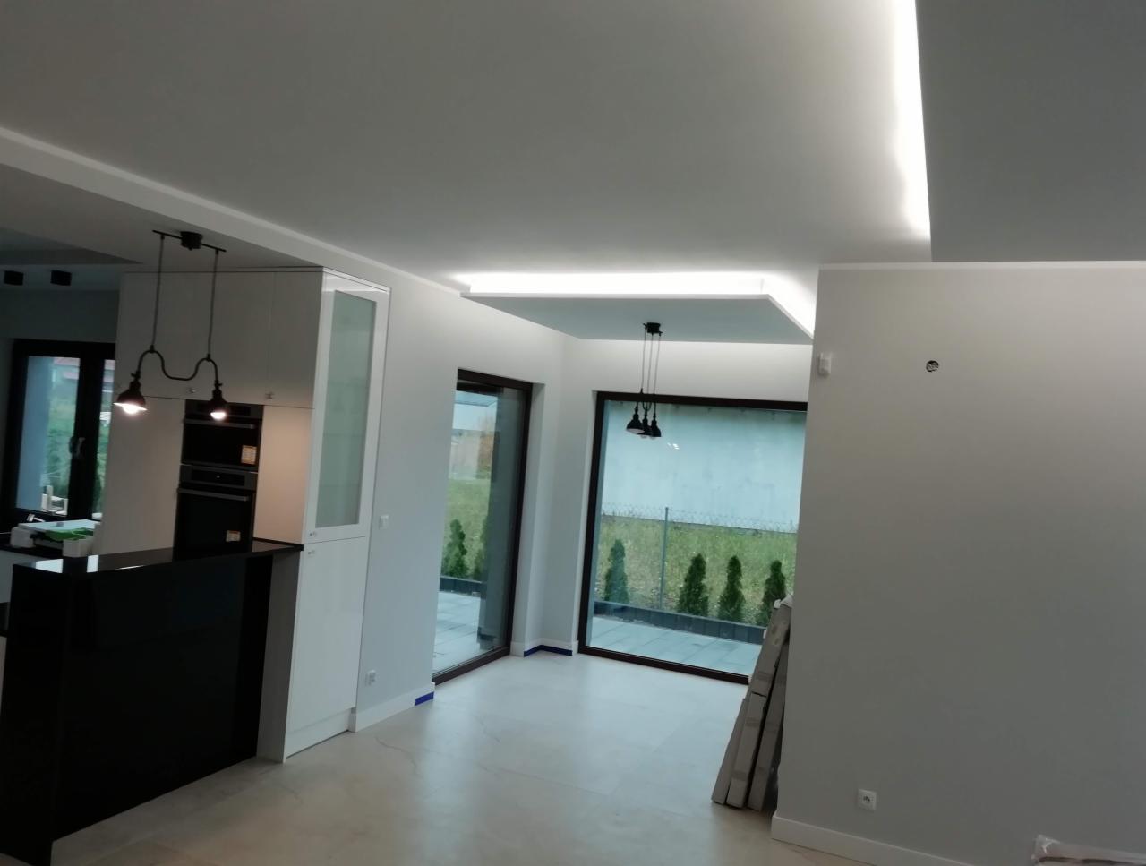 Nowoczesne wnętrze z oświetleniem LED w suficie podwieszanym, widoczne gładkie ściany, duże okna z widokiem na ogród i fragment zabudowy kuchennej z czarnym blatem.