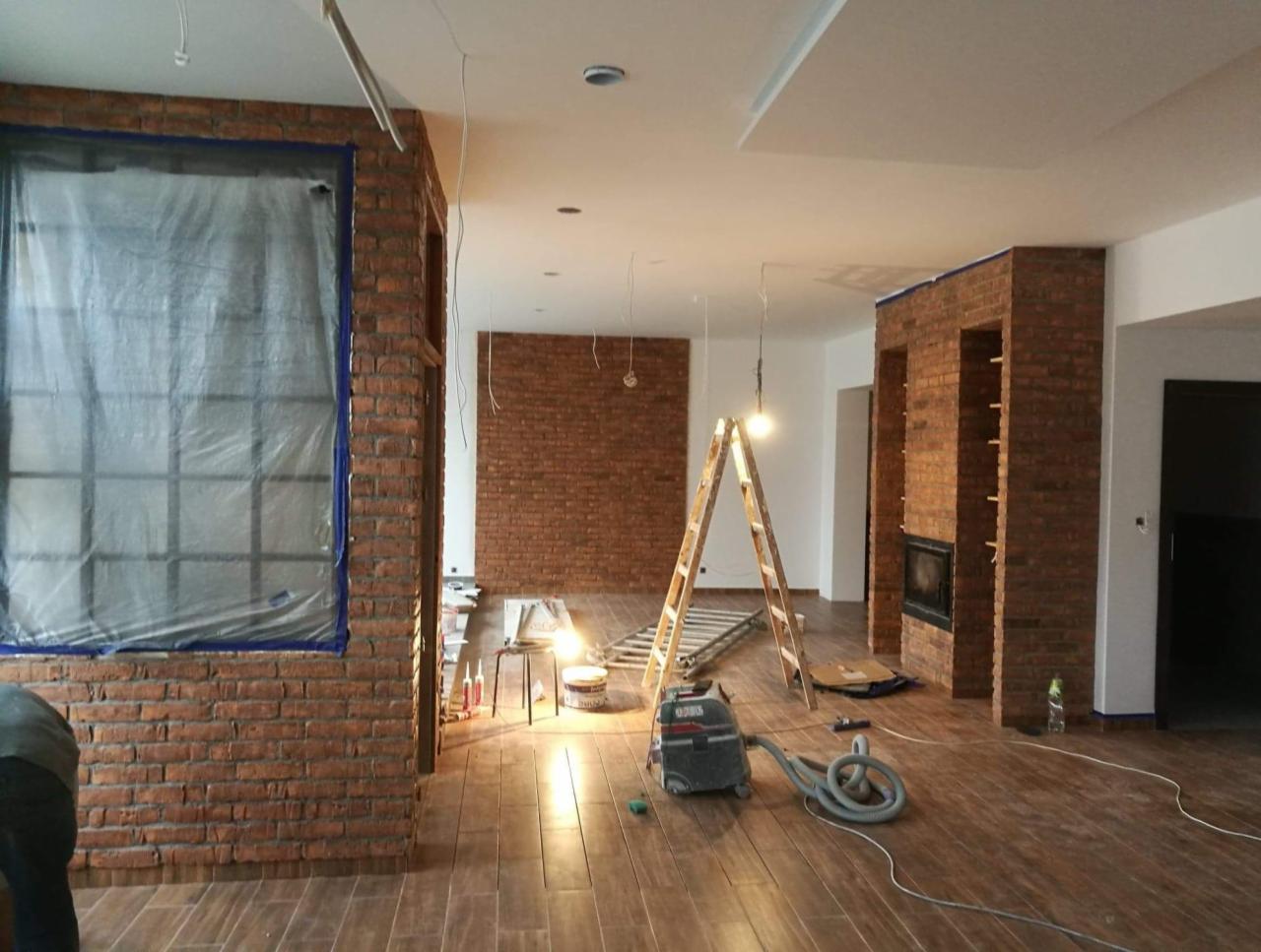Wnętrze w trakcie wykańczania z ceglaną ścianą, zabezpieczonym oknem folią malarską, drewnianą podłogą, drabiną i odkurzaczem przemysłowym.