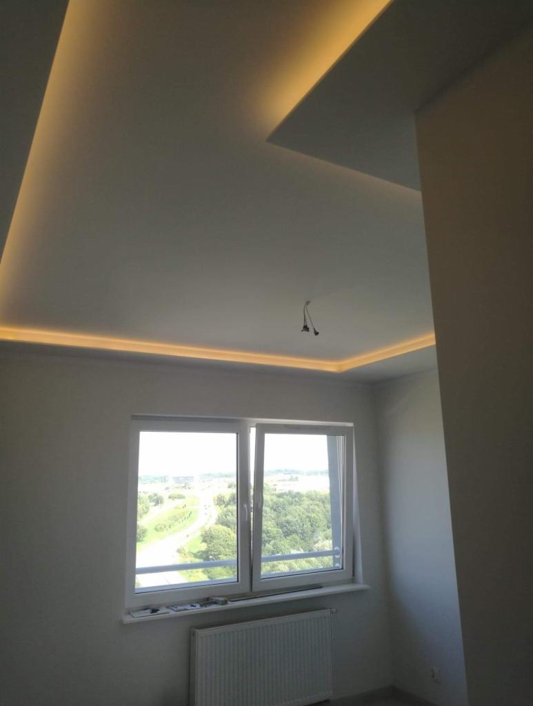 Nowoczesny pokój z białymi ścianami i podwieszanym sufitem z oświetleniem LED, widok z okna na zieleń za oknem.