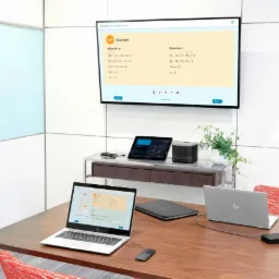 Sala konferencyjna z białym ekranem interaktywnym wyświetlającym zadania matematyczne, dwoma laptopami HP, tabletem, głośnikiem, białym panelem z notatkami i drewnianym stołem.