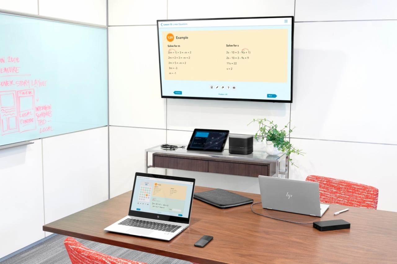 Sala konferencyjna z białym ekranem interaktywnym wyświetlającym zadania matematyczne, dwoma laptopami HP, tabletem, głośnikiem, białym panelem z notatkami i drewnianym stołem.