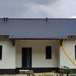 Nowo wybudowany parterowy dom z ciemną dachówką i jasną elewacją, widoczne żółte rury na zewnątrz, logo firmy na górze po prawej stronie.