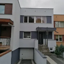 Dwukondygnacyjny dom z garażem pomalowany na biało i szaro, z widocznymi elementami dekoracyjnymi z cegły i szarą stolarką okienną, sfotografowany w pochmurny dzień.