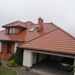 Dom jednorodzinny z nową, czerwoną dachówką karpiówką i ceglanym kominem, widok z góry w pochmurny dzień. W tle widoczne drzewa i część ogrodu z niebieskim wężem ogrodowym. Samochód osobowy...