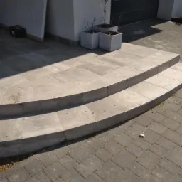 Dwu-stopniowe, półokrągłe schody z płytek ceramicznych prowadzące do domu, z trzema kwadratowymi donicami z roślinami przy wejściu i kostką brukową na podjeździe.