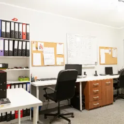 Jasne biuro rachunkowe z białymi ścianami, regałem pełnym segregatorów w kolorach czarnym, niebieskim i czerwonym, biurkami z komputerami, tablicą korkową z kalendarzem i tablicą suchościeralną...