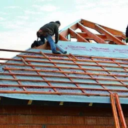 Dwóch dekarzy pracuje na dachu domu w trakcie budowy, układając drewniane łaty na membranie dachowej, widoczna więźba dachowa i fragment murowanej ściany.