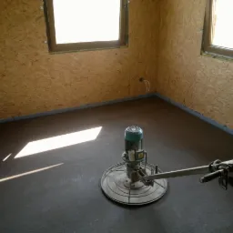 Zacieranie posadzki jastrychowej w pomieszczeniu z ścianami wyłożonymi płytą OSB. Maszyna do zacierania posadzek na świeżo wylanej, szarej posadzce. Widoczne okna.