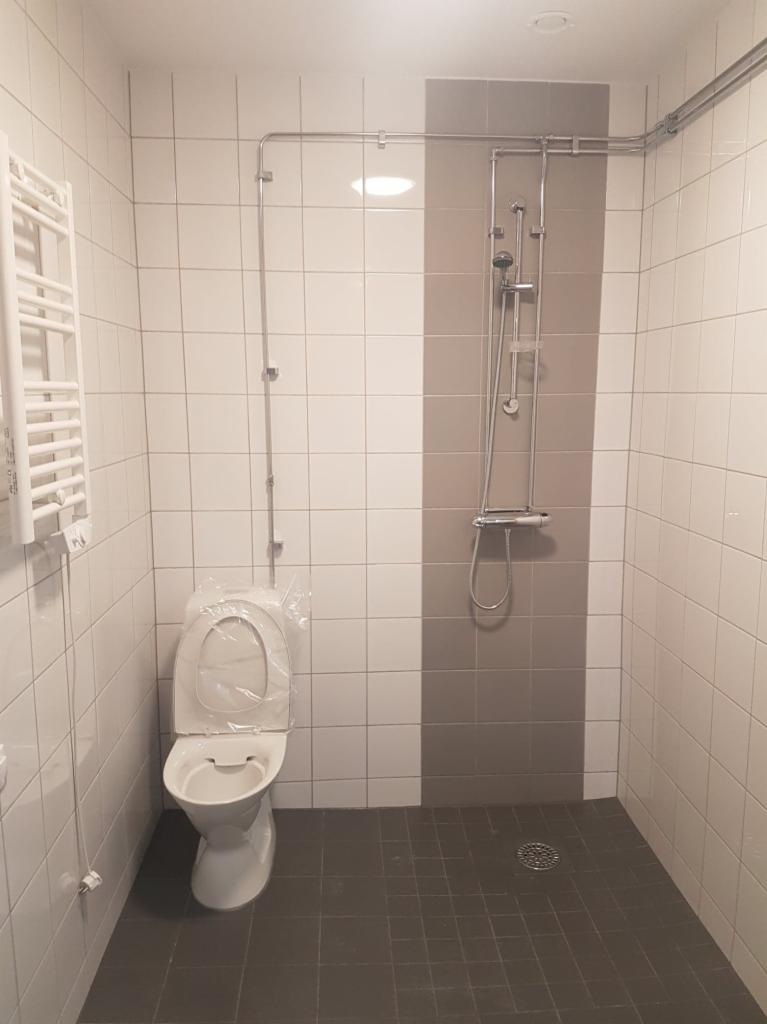 Wnętrze wykończonej łazienki z biało-szarą glazurą, nową toaletą zabezpieczoną folią i zamontowanym zestawem prysznicowym z widoczną instalacją hydrauliczną.