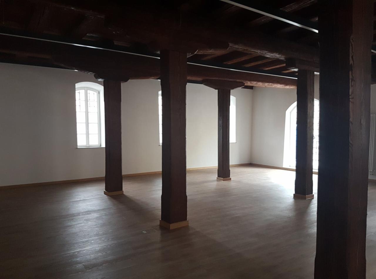 Wnętrze przestronnego pomieszczenia z drewnianą podłogą, masywnymi, ciemnymi drewnianymi słupami i belkami stropowymi, jasnymi ścianami i wysokimi oknami.