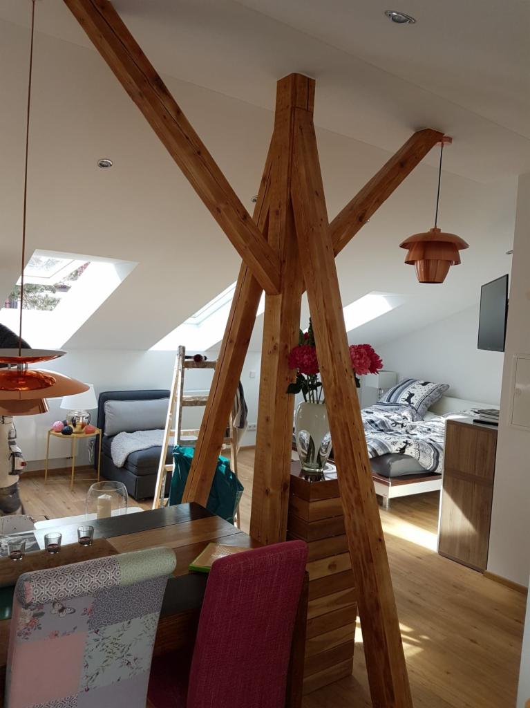 Jasne poddasze z drewnianą konstrukcją nośną, widoczne okna dachowe, łóżko z wzorzystą pościelą, drewniana drabina oparta o ścianę, lampa wisząca w stylu retro i stół z krzesłami.