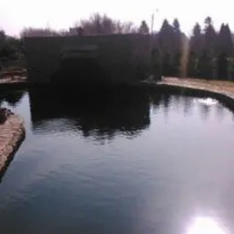 Duże oczko wodne z kamienną obudową i kaskadą, otoczone brukowanym chodnikiem i zielenią, odbicie słońca na powierzchni wody.