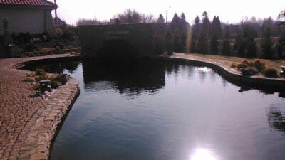 Duże oczko wodne z kamienną obudową i kaskadą, otoczone brukowanym chodnikiem i zielenią, odbicie słońca na powierzchni wody.