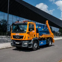 Pomarańczowo-niebieska ciężar&oacute;wka hakowa MAN z kontenerem na gruz firmy Mustang-Gliwice na tle nowoczesnego budynku z ciemną fasadą i szklanymi panelami.