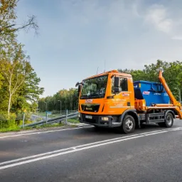 Pomarańczowa ciężarówka z niebieskim kontenerem na platformie, jadąca po asfaltowej drodze z podwójną linią ciągłą w słoneczny dzień, z zielonymi drzewami w tle.