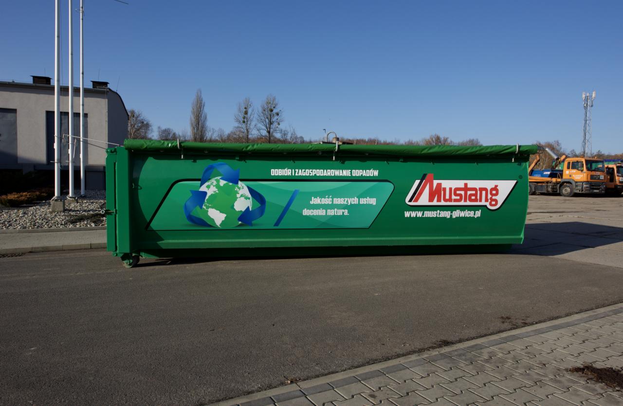 Zielony kontener na odpady firmy Mustang z logo i hasłem 'Odbiór i zagospodarowanie odpadów' na tle parkingu i błękitnego nieba.