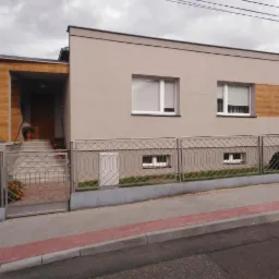 Nowoczesny dom jednorodzinny z jasnoszarą elewacją, drewnianym akcentem na ścianie i przy wejściu, ogrodzony metalowym płotem, widok z poziomu ulicy w pochmurny dzień.