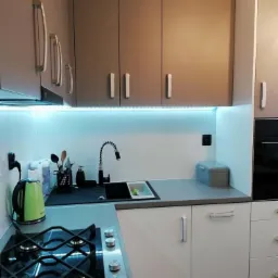 Nowoczesna kuchnia z białymi i beżowymi szafkami, podświetleniem LED pod górnymi szafkami, czarnym zlewem i płytą gazową na szarym blacie.