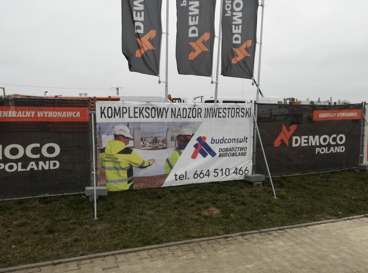Baner reklamowy z napisem 'Kompleksowy Nadzór Inwestorski' i logotypem firmy budowlanej, umieszczony na ogrodzeniu placu budowy z widocznymi maszynami budowlanymi w tle.