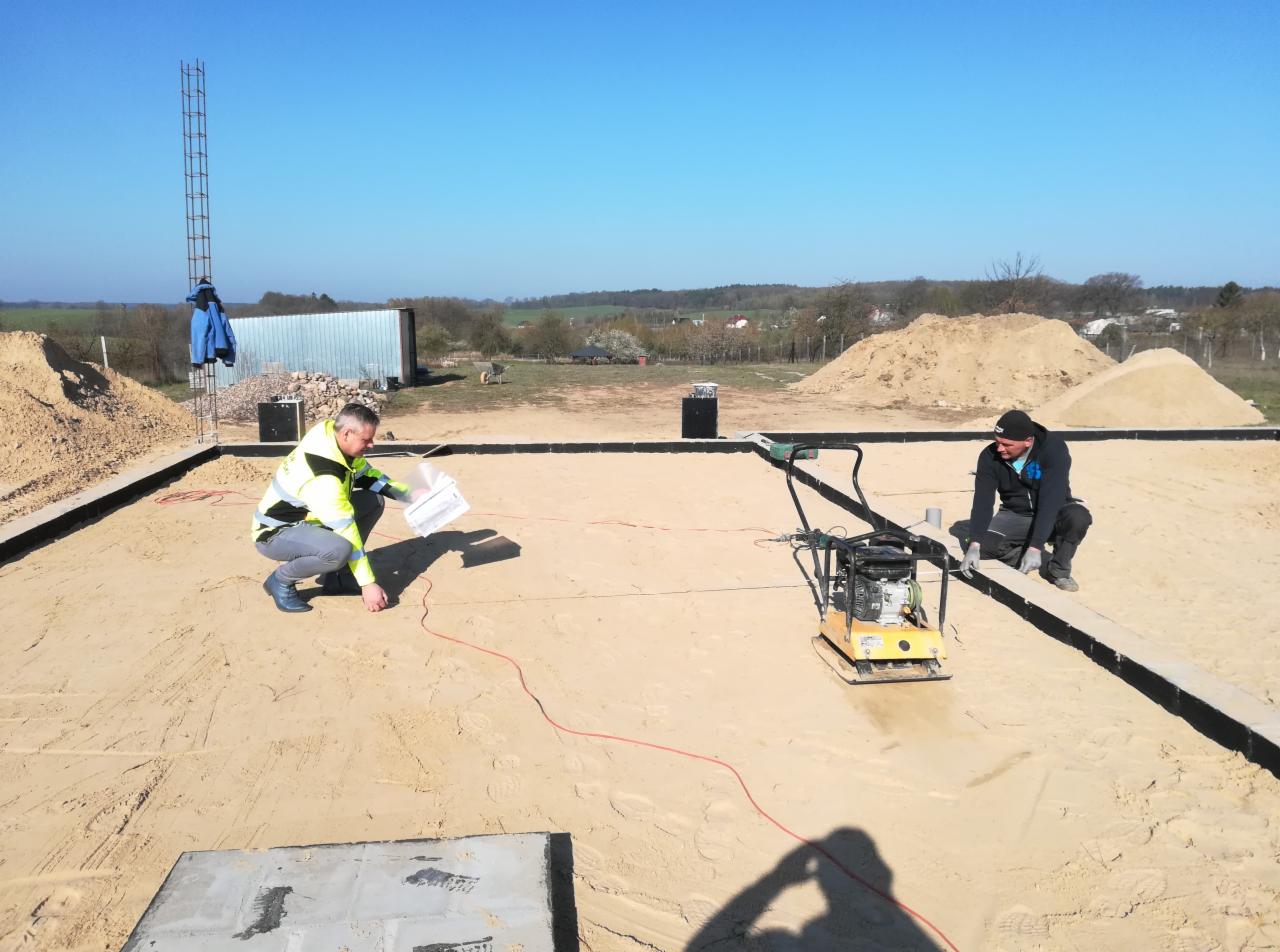 Dwóch pracowników na placu budowy wyrównuje piasek zagęszczarką, jeden sprawdza dokumentację, widoczne zbrojenie, hałdy piasku i krajobraz