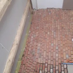 Ceglana podłoga ułożona w jodełkę, widoczne deski stropowe i płyty gipsowo-kartonowe na ścianach, narzędzia budowlane w rogu, etap prac wykończeniowych.