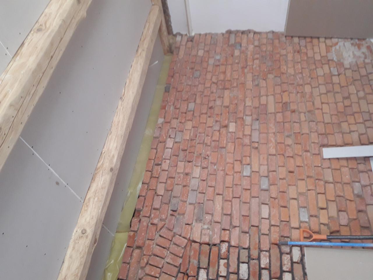 Ceglana podłoga ułożona w jodełkę, widoczne deski stropowe i płyty gipsowo-kartonowe na ścianach, narzędzia budowlane w rogu, etap prac wykończeniowych.