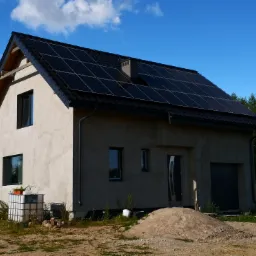 Dom jednorodzinny z zamontowanymi panelami fotowoltaicznymi na dachu, widok z zewnątrz, na pierwszym planie pryzma piachu.