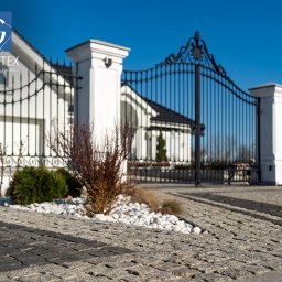 PRZEDSIĘBIORSTWO PRODUKCYJNO HANDLOWO USŁUGOWE „GRANITEX” RESIN SYSTEMS SŁAWOMIR MENDEL - Granitowa kostka brukowa w Sławnie, ułożona przed posesją z białym domem i ozdobnym ogrodzeniem. Widoczny fragment trawnika i dekoracyjne kamienie.