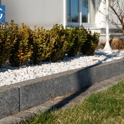PRZEDSIĘBIORSTWO PRODUKCYJNO HANDLOWO USŁUGOWE „GRANITEX” RESIN SYSTEMS SŁAWOMIR MENDEL - Granitowe obrzeże rabaty z białymi kamykami, oddzielające trawnik od krzewów, w tle ściana budynku z oknem, oświetlone słońcem.