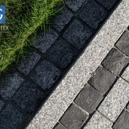 PRZEDSIĘBIORSTWO PRODUKCYJNO HANDLOWO USŁUGOWE „GRANITEX” RESIN SYSTEMS SŁAWOMIR MENDEL - Ułożona kostka granitowa w dwóch odcieniach szarości, oddzielona pasem jaśniejszego granitu, z fragmentem trawy w rogu. Detal nawierzchni.