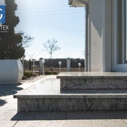 PRZEDSIĘBIORSTWO PRODUKCYJNO HANDLOWO USŁUGOWE „GRANITEX” RESIN SYSTEMS SŁAWOMIR MENDEL - Granitowe schody wejściowe do budynku, z widoczną kostką granitową na podłożu i elementami architektury ogrodowej w tle. Jasne, słoneczne oświetlenie.