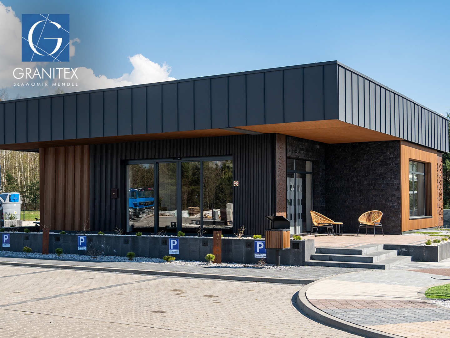 Nowoczesny showroom GRANITEX z elementami drewnopodobnymi, kostką brukową i granitową na zewnątrz. Widoczne miejsca parkingowe i detale wykończenia.
