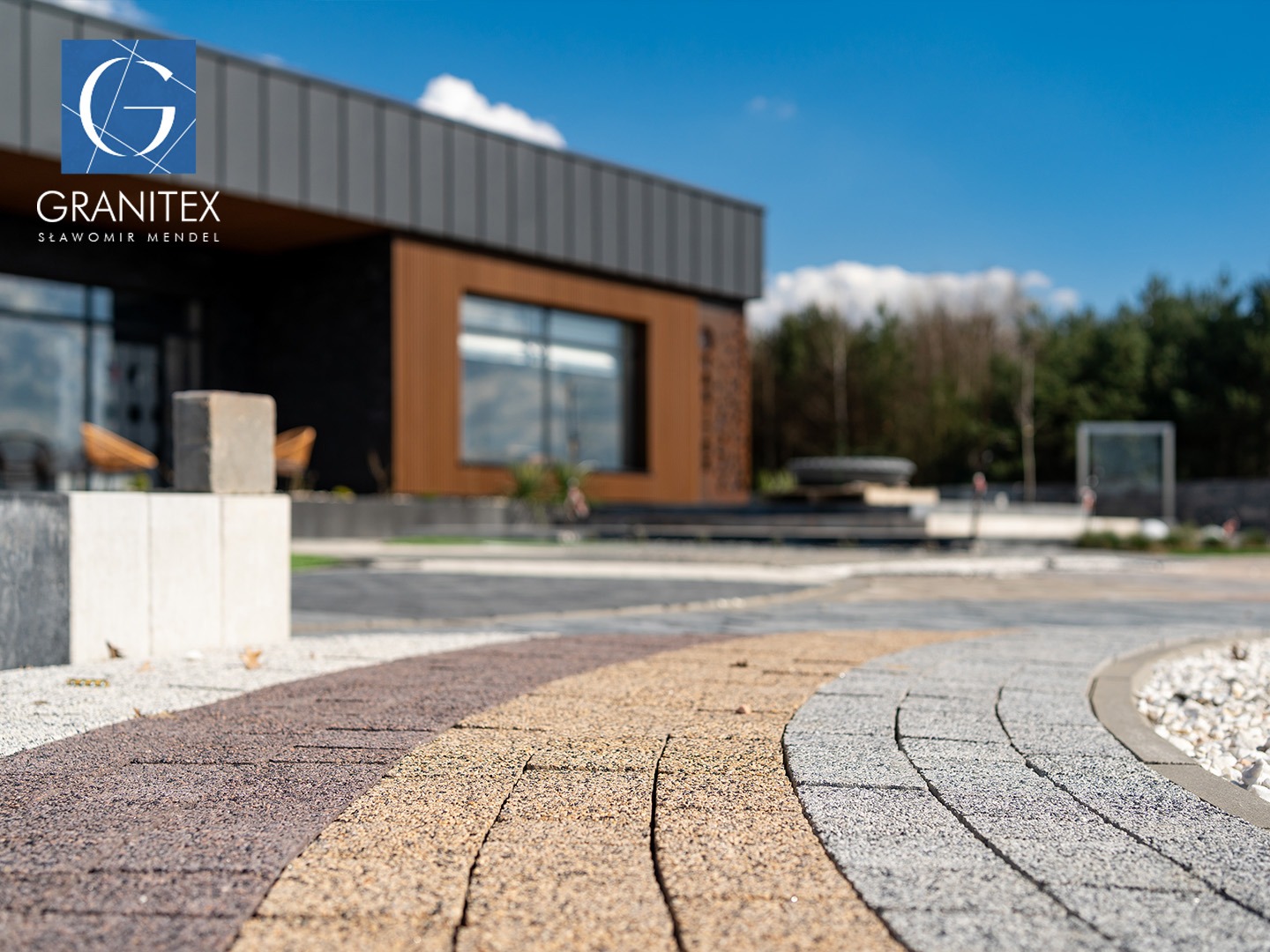 Wzorcowa ścieżka z kostki brukowej w odcieniach brązu, beżu i szarości, prowadząca do nowoczesnego budynku z logo Granitex na tle błękitnego nieba.