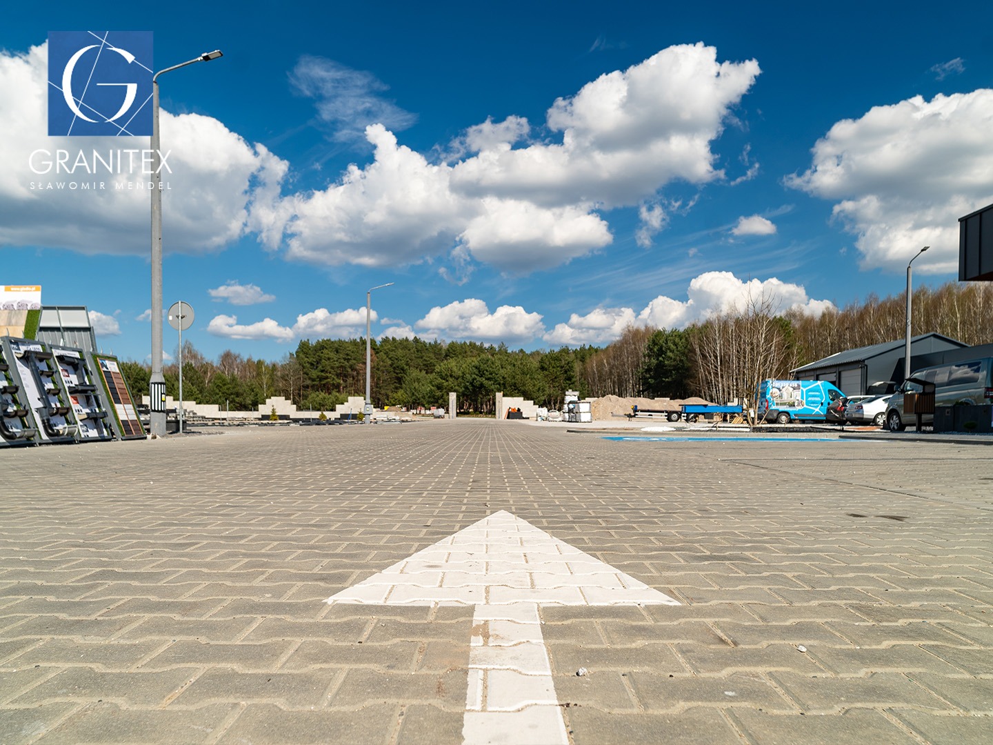 Rozległy plac wyłożony szarą kostką brukową z białą strzałką kierunkową. W tle ekspozycja kostki, zieleń, budynek i firmowe samochody w słoneczny dzień.