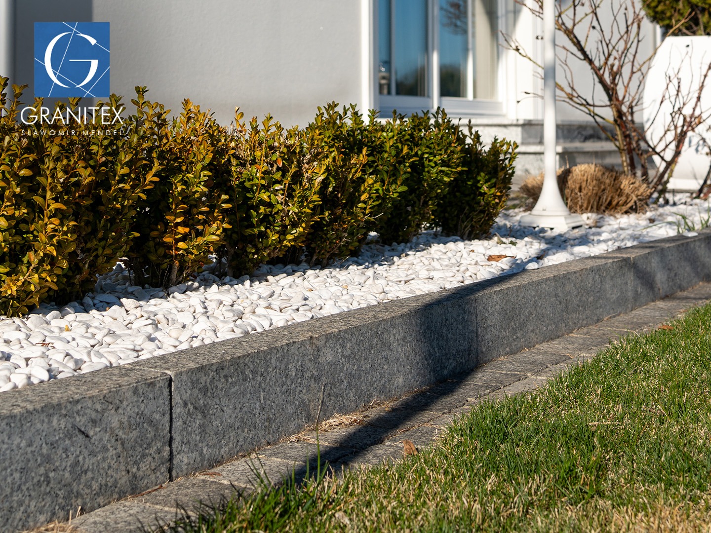 Granitowe obrzeże rabaty z białymi kamykami, oddzielające trawnik od krzewów, w tle ściana budynku z oknem, oświetlone słońcem.