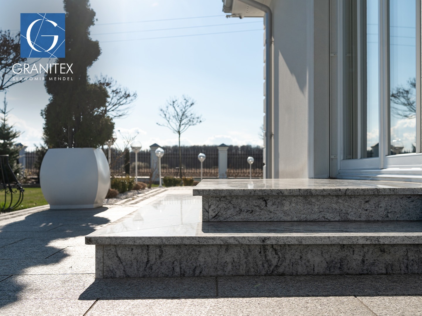 Granitowe schody wejściowe do budynku, z widoczną kostką granitową na podłożu i elementami architektury ogrodowej w tle. Jasne, słoneczne oświetlenie.
