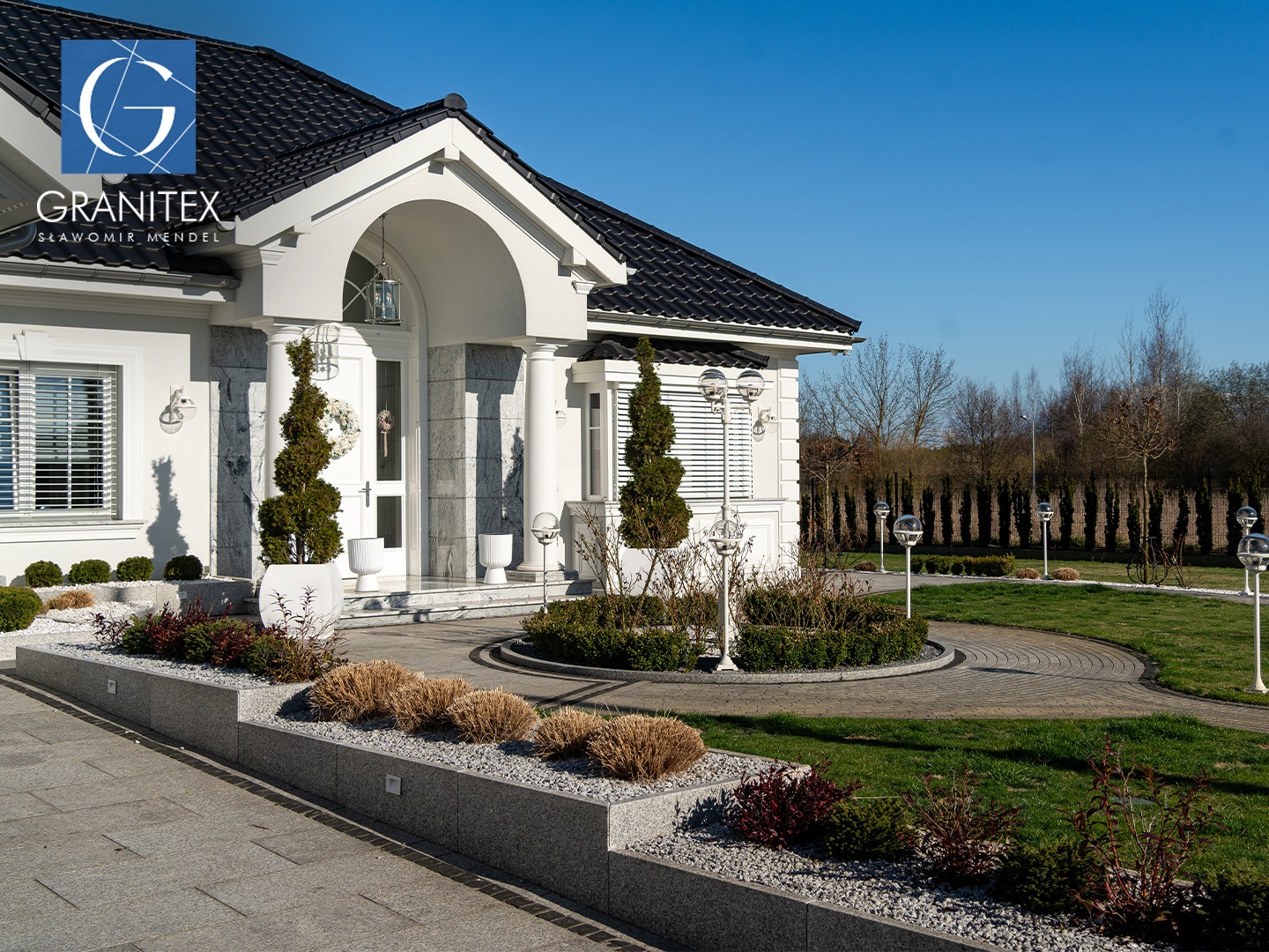 Elegancki dom z granitową elewacją i brukowanym podjazdem. Ozdobne rośliny i lampy kuliste podkreślają estetykę posesji.