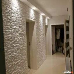 Korytarz z dekoracyjną ścianą wyłożoną białymi panelami imitującymi kamień, oświetlony punktowymi lampami sufitowymi. Widoczne białe drzwi i fragment otwartej szafy wnękowej w tle.