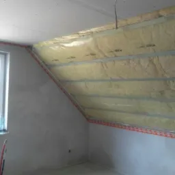 Poddasze w trakcie ocieplania wełną mineralną, widoczne profile montażowe, folia paroizolacyjna i surowy tynk na ścianach, okno z butelką wody na parapecie.