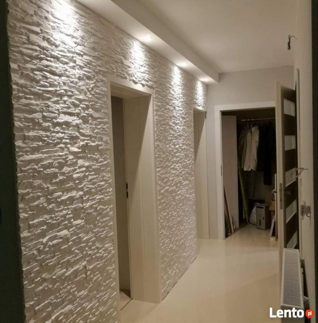 Korytarz z dekoracyjną ścianą wyłożoną białymi panelami imitującymi kamień, oświetlony punktowymi lampami sufitowymi. Widoczne białe drzwi i fragment otwartej szafy wnękowej w tle.