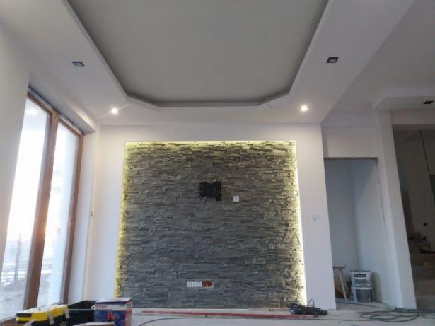 Salon z oświetloną ścianą wyłożoną dekoracyjnym kamieniem, przygotowany pod montaż telewizora, widoczne narzędzia i materiały budowlane.