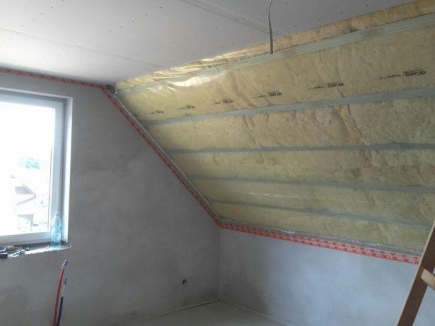Poddasze w trakcie ocieplania wełną mineralną, widoczne profile montażowe, folia paroizolacyjna i surowy tynk na ścianach, okno z butelką wody na parapecie.