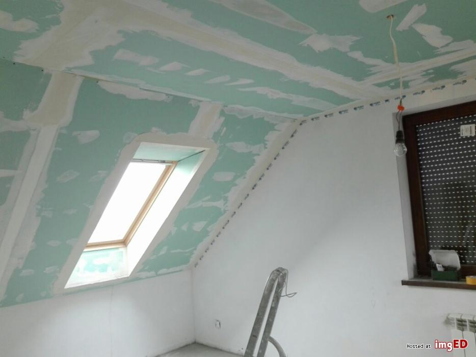 Montaż płyt gipsowo-kartonowych na poddaszu ze skosami i oknem dachowym, widoczne łączenia płyt przed szpachlowaniem, surowa żarówka zwisająca z sufitu, drabina oparta o ścianę.