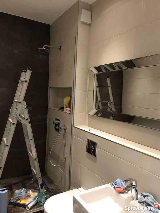 Wnętrze łazienki w trakcie remontu; widoczna drabina oparta o ścianę z prysznicem, umywalka z baterią i lustro z ozdobną ramą; na podłodze narzędzia i środki czystości.