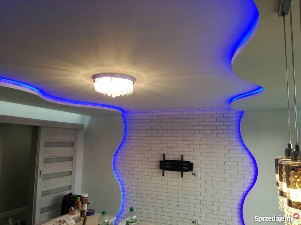 Sufit podwieszany z nieregularnymi kształtami, podświetlony niebieską listwą LED, ściana z białej cegły z uchwytem na telewizor, kryształowy żyrandol w centralnej części sufitu.