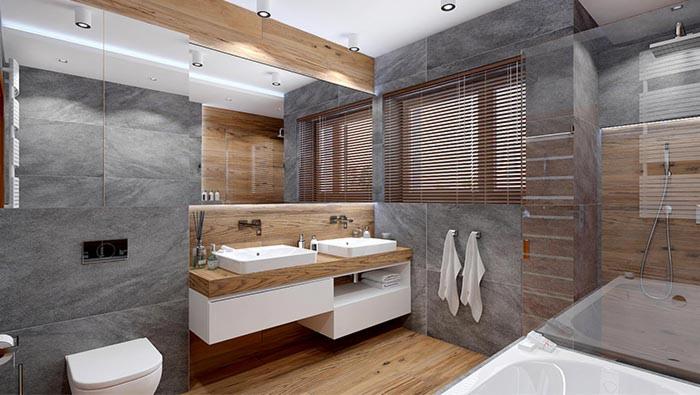 Nowoczesna łazienka z szarymi płytkami imitującymi kamień, drewnianymi akcentami, podwójną umywalką na białej szafce, lustrem z drewnianą ramą i oknem z żaluzjami.