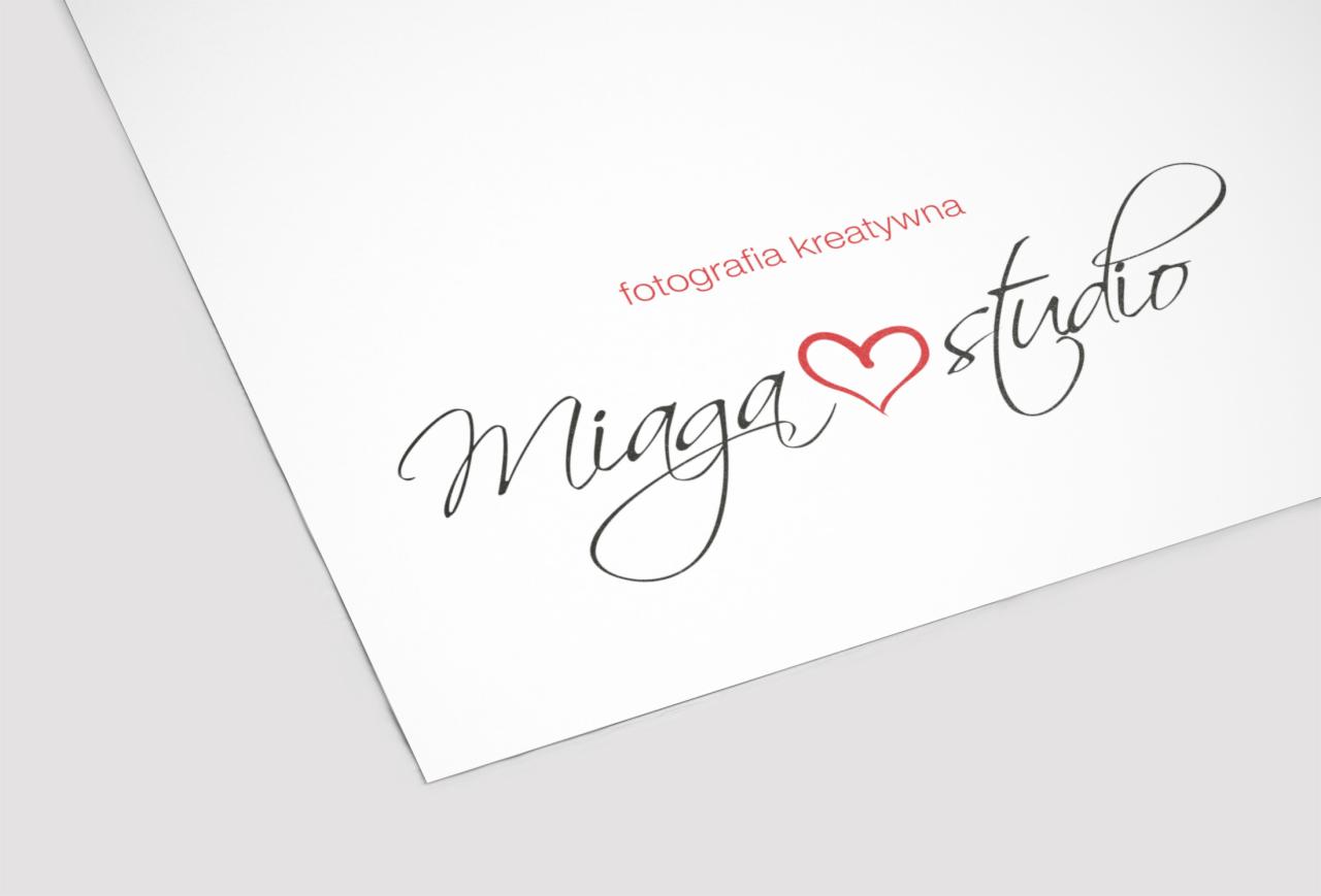 Logo studia fotograficznego 'Miaga studio' z czerwonym sercem, umieszczone na białym arkuszu papieru, delikatnie pochylonym i oświetlonym z góry.