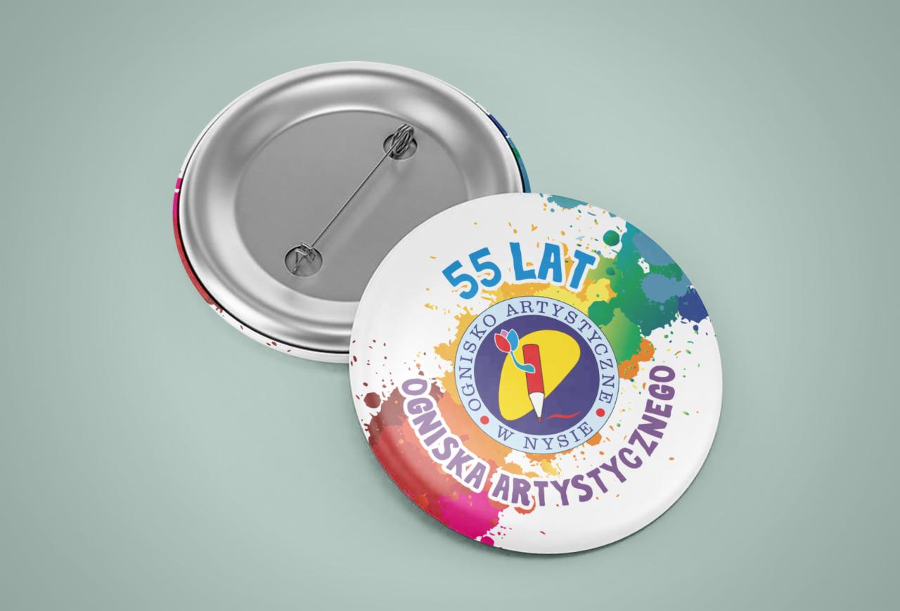 Dwie okrągłe przypinki. Jedna z metalowym tyłem i agrafką, druga z kolorowym nadrukiem logo 'Ogniska Artystycznego w Nysie' z okazji 55-lecia, otoczonym plamami farb w różnych kolorach.